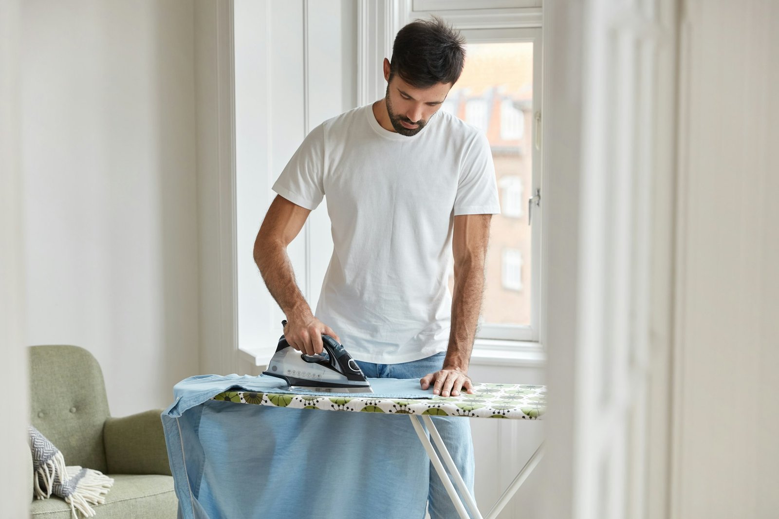 Photo of busy unshaven man irons shirt on ironing board, prepares for formal meeting on business con