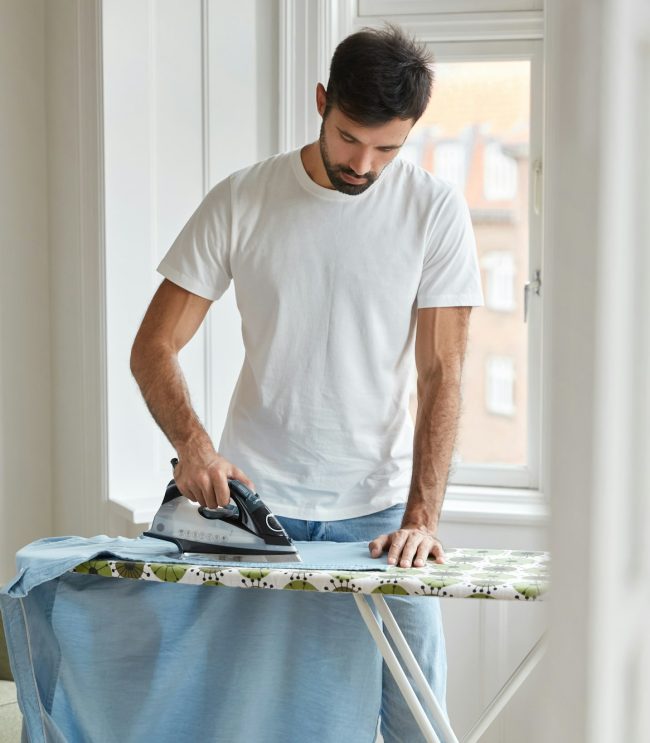 Photo of busy unshaven man irons shirt on ironing board, prepares for formal meeting on business con Photo of busy unshaven man irons shirt on ironing board, prepares for formal meeting on business con